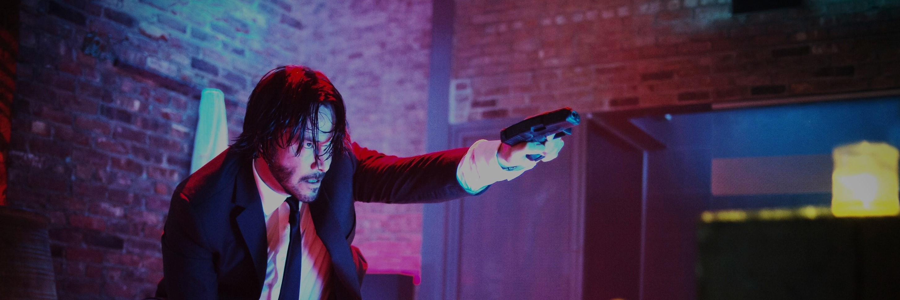 a man called John Wick, dressed in a black suit with a white button up shirt and a black tie, holding a gun and aiming away to the right looking very intensly at his target.