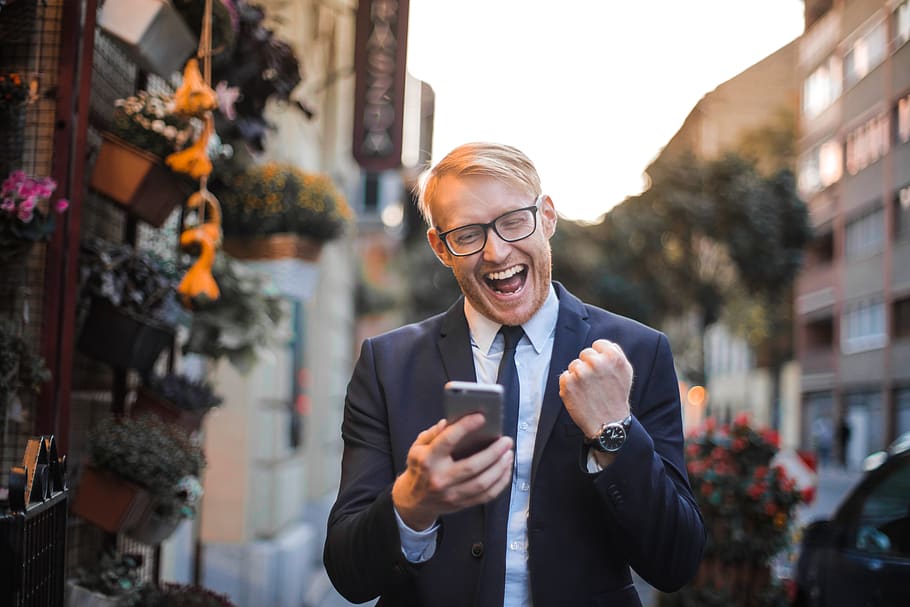 A buisness man holding a phone and smiling happily and looking overall excited over his phone.